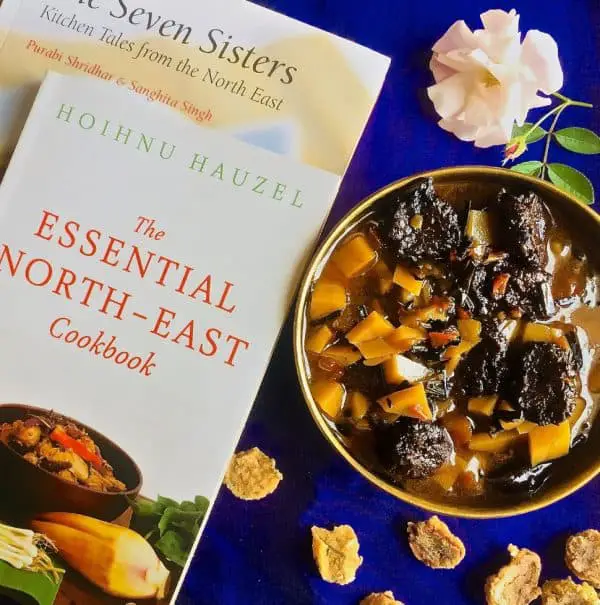 970×978 Aloo Badi A brass bowl with aloo badi, a Manipuri dish of potatoes and little sundried nuggets of black gram lentil paste, on a purple background and book on the North East Indian cuisine alongside