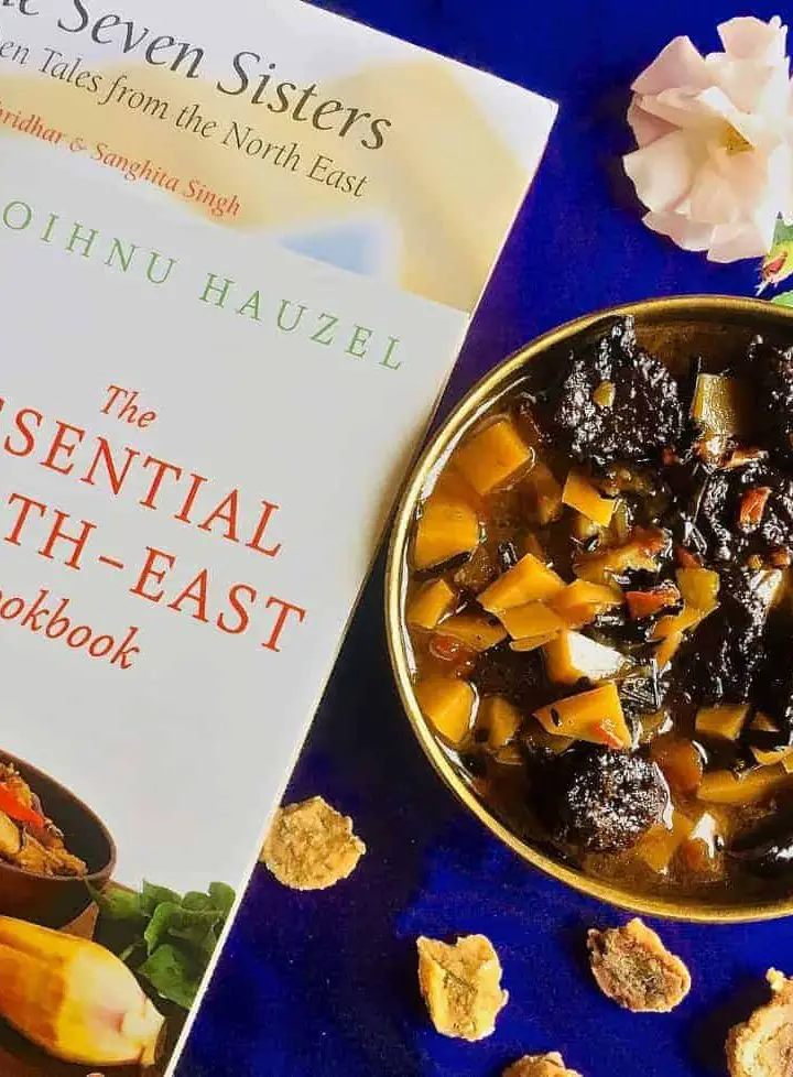 A brass bowl with aloo badi, a Manipuri dish of potatoes and little sundried nuggets of black gram lentil paste, on a purple background and book on the North East Indian cuisine alongside