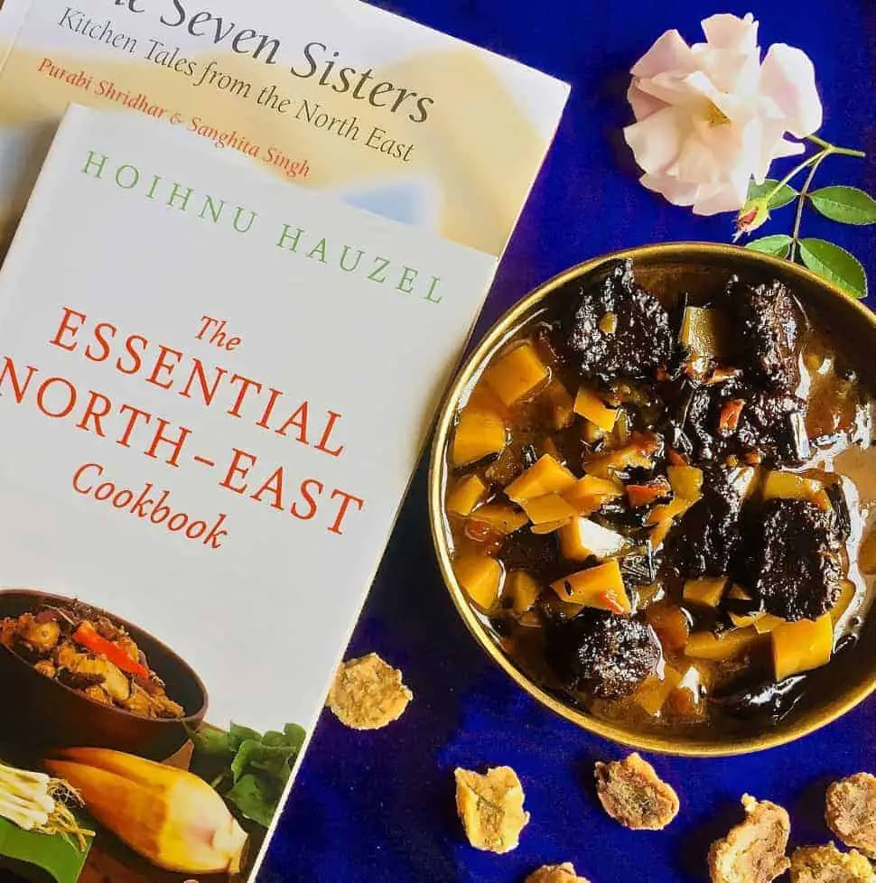 A brass bowl with aloo badi, a Manipuri dish of potatoes and little sundried nuggets of black gram lentil paste, on a purple background and book on the North East Indian cuisine alongside