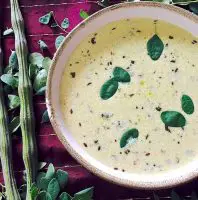 A bowl of murungai keerai morekuzhambu on a red silk cloth with 2 drumsticks and moringa leaves on the left side