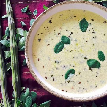 A bowl of murungai keerai morekuzhambu on a red silk cloth with 2 drumsticks and moringa leaves on the left side