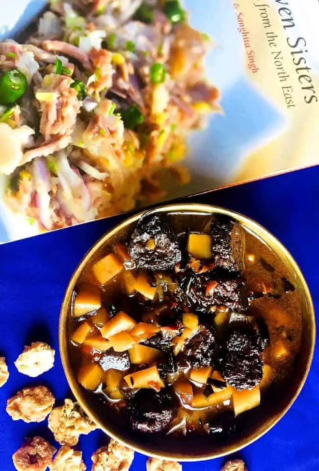 A bowl of curry of potato, chives and fried lentil nuggets on a dark blue background with book on North East Indian cusine above