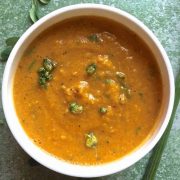 A white bowl with mustard orange coloured Drumstick Corn Carrot Soup with Superfood Moringa leaves and pods. Garnished with moringa flowers. On a green background with a green moringa pod on one side and a few moringa leaves scattered on the other side