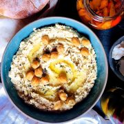 A large shallow blue bowl of creamy yellow hummus with olive oil and chickpeas for garnish, with a plate of pita bread, a jar of roasted red peppers, bowl of goats cheese and jar of olives surrounding