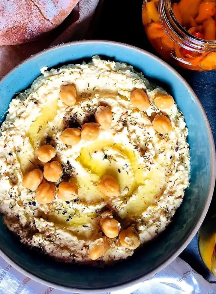 A large shallow blue bowl of creamy yellow hummus with olive oil and chickpeas for garnish, with a plate of pita bread, a jar of roasted red peppers, bowl of goats cheese and jar of olives surrounding