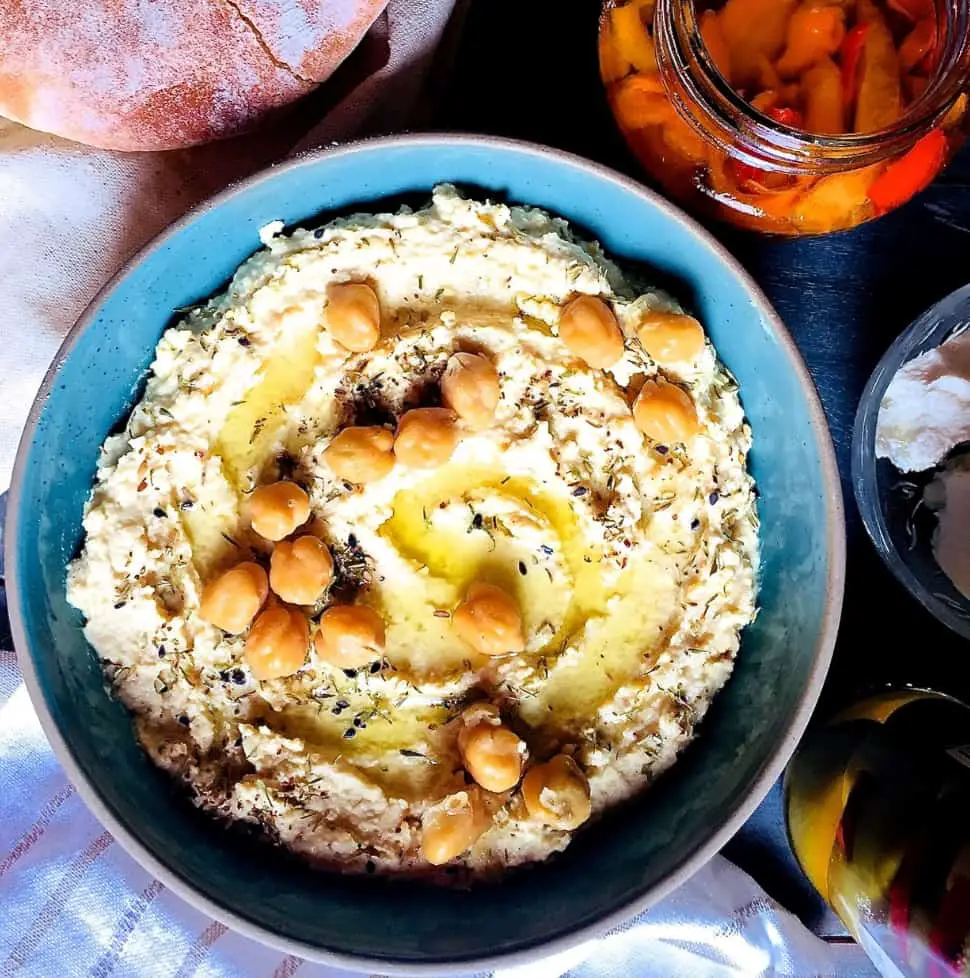 A large shallow blue bowl of creamy yellow hummus with olive oil and chickpeas for garnish, with a plate of pita bread, a jar of roasted red peppers, bowl of goats cheese and jar of olives surrounding