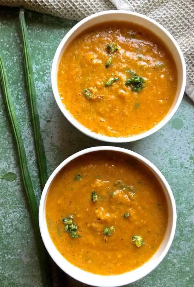 Two white soup bowls with orangey drumstick corn carrot soup. Garnished with moringa flowers and leaves and moringa pods seen to the left