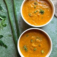 Two white bowls with mustard orange coloured Drumstick Corn Carrot Soup with Superfood Moringa leaves and pods. Garnished with moringa flowers. On a green background with green moringa pods and a few moringa leaves scattered on the side