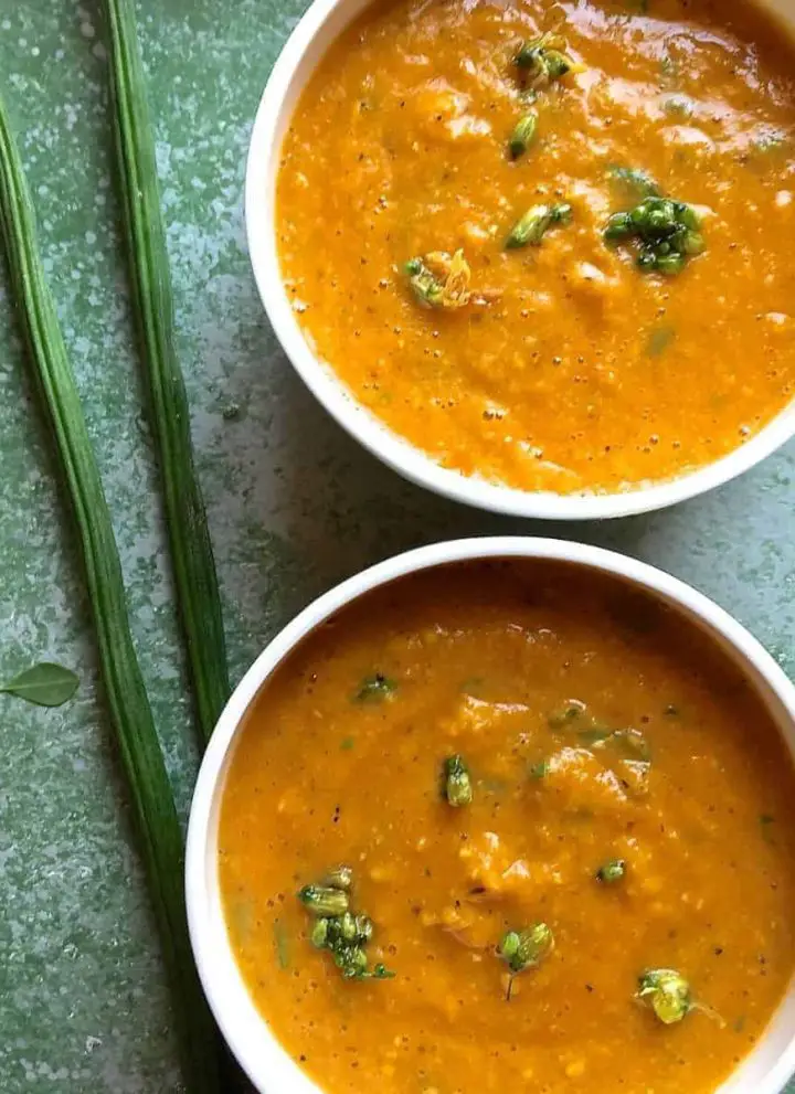 Two white bowls with mustard orange coloured Drumstick Corn Carrot Soup with Superfood Moringa leaves and pods. Garnished with moringa flowers. On a green background with green moringa pods and a few moringa leaves scattered on the side