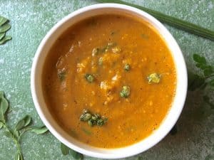 A large white bowl filled with orange mustard moringa pods leaves carrot and corn soup. On a light green background. with a drumstick above and moringa leaves alongside. Garnsihed with fried moringa blossoms