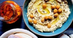 A wide blue bowl of a pale creamy yellow hummus, with chickpeas and olive oil for garnish and with a plate of pita bread and a jar of roasted red peppers alongside