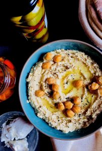 A large shallow blue dish spread with hummus, garnished with chickpeas and za'atar spice, Olive oil drizzled across its surface. A jar of roasted red peppers in oil on one side and a jar of stuffed olives in another