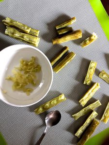 A white bowl with some scopped out moringa seeds. A few moringa drumstick pieces scattered on the grey background. Some sliced into two vertically, showing the seeds, some empty after the seeds have been scopped out with a spoon