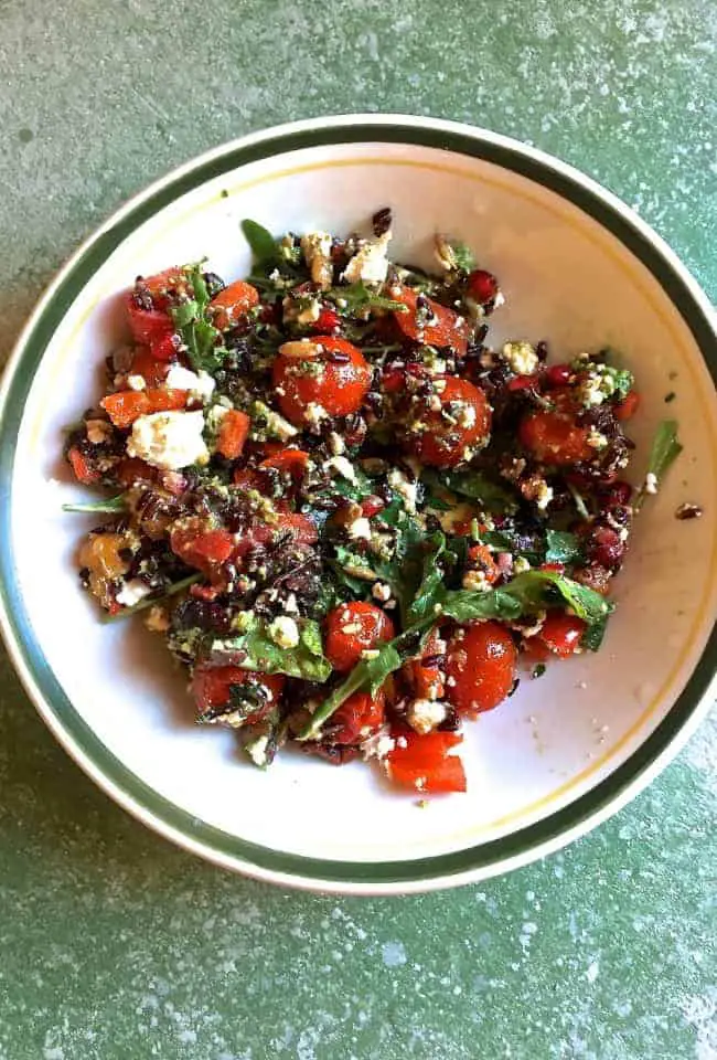 A green edged white plate with black rice, red and yellow cherry tomatoes, greens, goat cheese and seeds in a parsley pesto