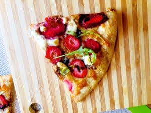 A wedge shaped slice of strawberry goat cheese pizza with arugula leaves and balsamic reduction, on a stilped wooden board