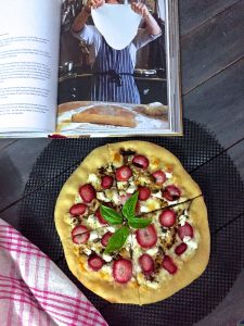 A book with a recipe and image of pizza being made, along with strawberry goat cheese pizza baked and shown with red striped napking