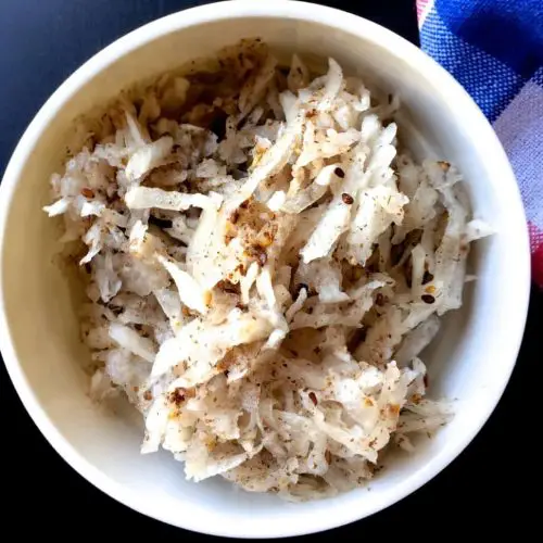 Large white bowl of grated white radish mixed with a brown paste, on dark blue black background. Black Sesame Radish salad from India's N E Meghalaya