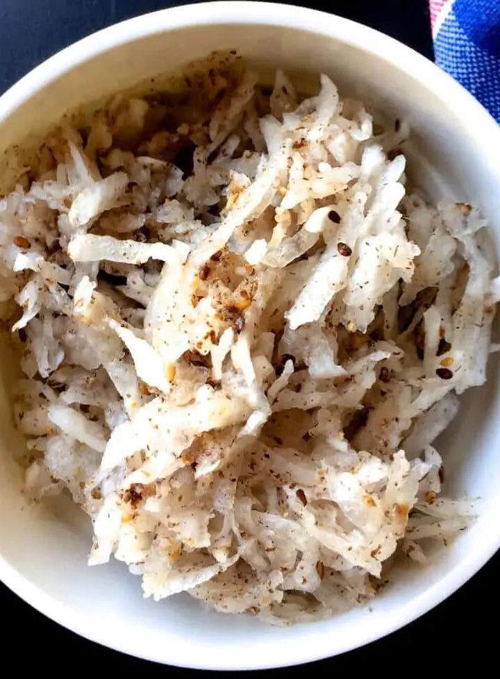 Large white bowl of grated white radish mixed with a brown paste, on dark blue black background. Black Sesame Radish salad from India's N E Meghalaya