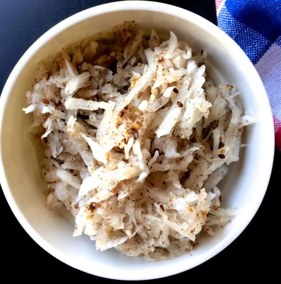 Large white bowl of grated white radish mixed with a brown paste, on dark blue black background. Black Sesame Radish salad from India's N E Meghalaya