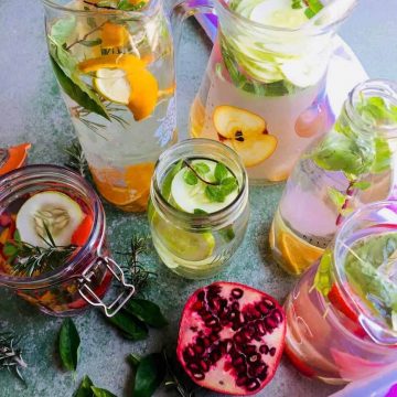 Glass Jars of flavoured water, infused with citrus fruit, cucumber, stawberries with basil, rosemary or mint herbs, and a sliced pomegranate, on a pale green background