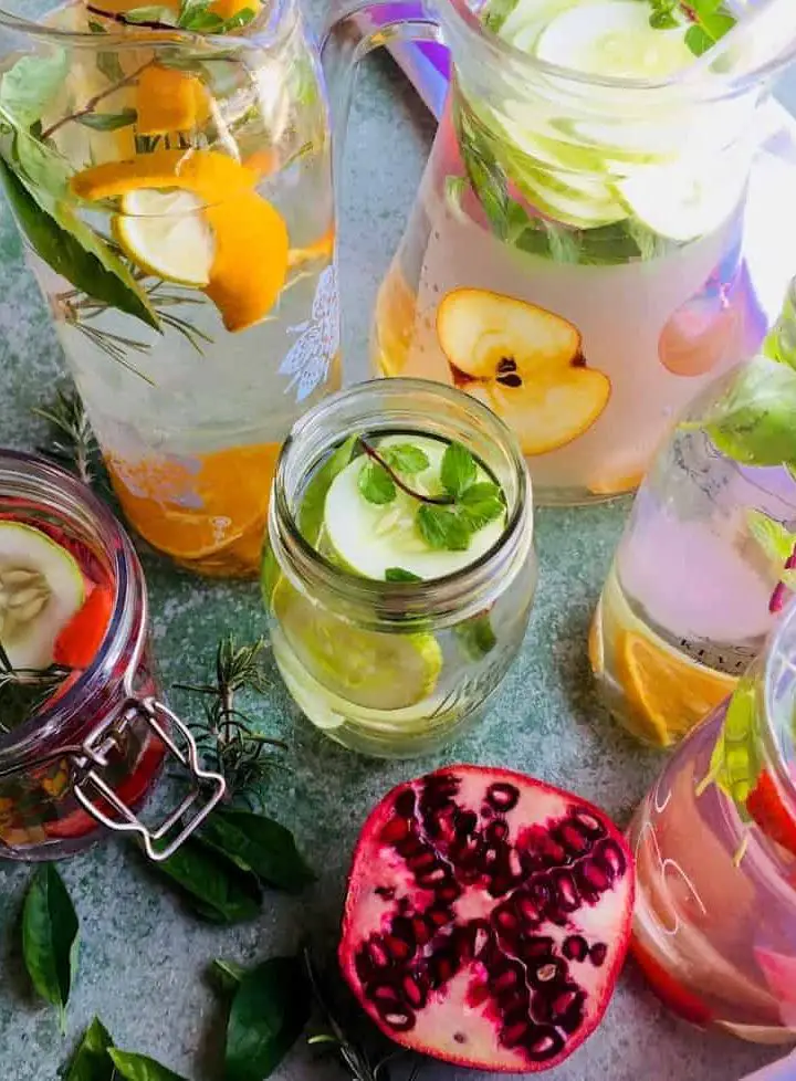 Glass Jars of flavoured water, infused with citrus fruit, cucumber, strawberries with basil, rosemary or mint herbs, and a sliced pomegranate, on a pale green background