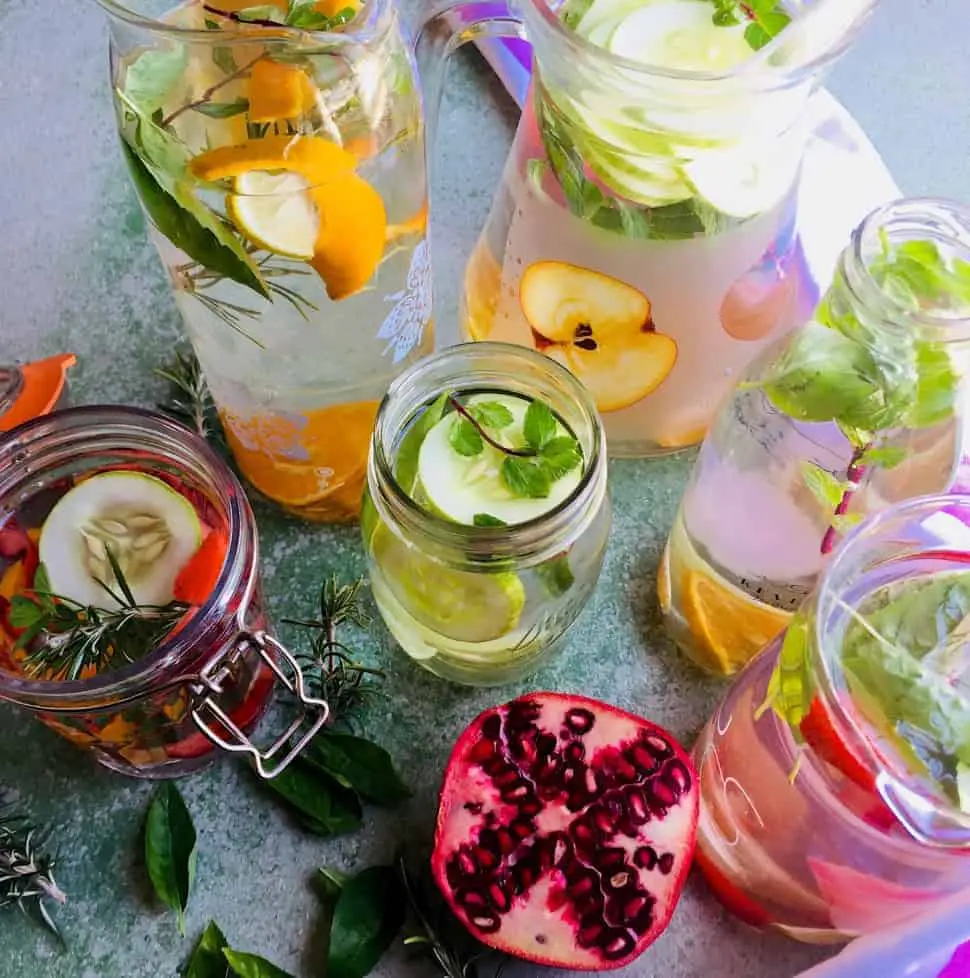 Glass Jars of flavoured water, infused with citrus fruit, cucumber, strawberries with basil, rosemary or mint herbs, and a sliced pomegranate, on a pale green background