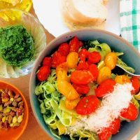 A bowl of fettucine pasta slathered with green pesto and red and yellowburst cherry tomatoes heaped on top with grated parmesan cheese. Bowls of pistachio and argula pesto on the side, with a bottle of olive oil and slices of bread