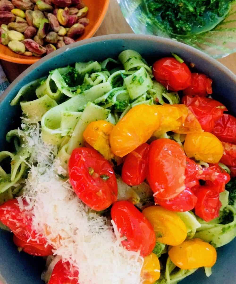 Fettucine pasta in green pesto with cherry tomato sauce heaped on top with grated parmesan. Bowl of pista and a bowl of pesto seen in the background