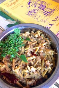 A large bowl filled with Bamboo shoot curry with coriander and curry leaves heaped on top to the left of the bowl. A yellow coloured book set in a fictional town of Malgudi in Karnataka, on one side