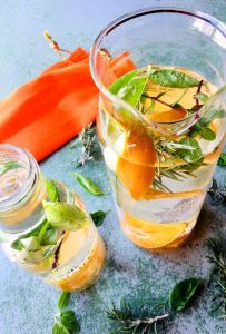 A tall glass jar and a smaller one with orange and lemon peel and slices, basil and mint and an orange fabric in the background to bring out the citrussy colours