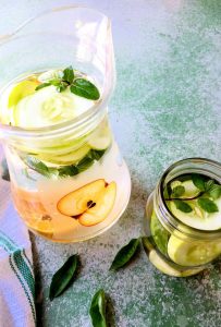 A tall glass jug of water with round slices of cucumber some lemon and sprigs of mint and a smaller jar with cucumber and mint