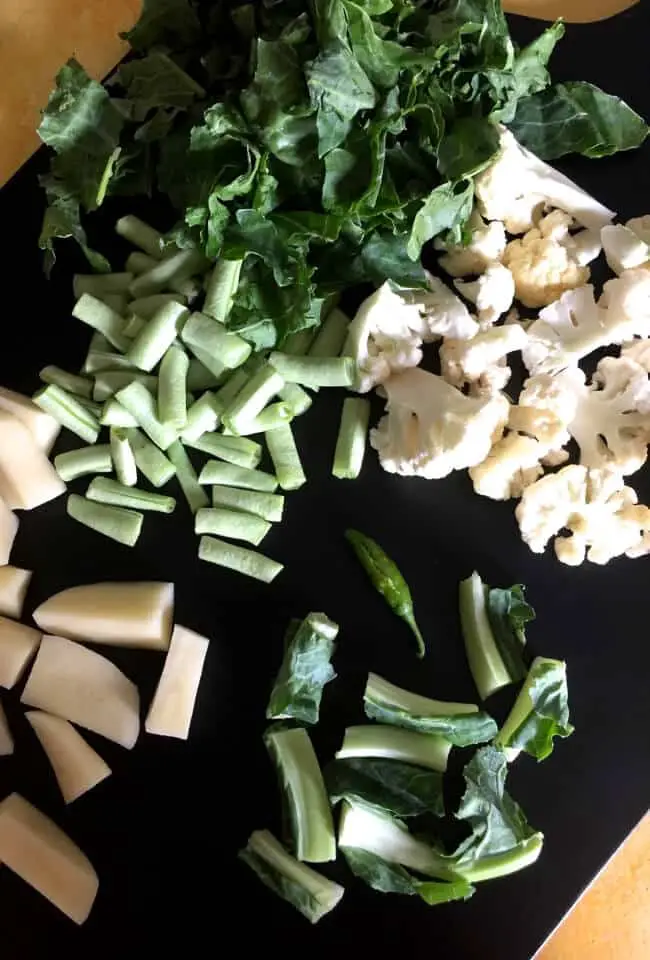 Ingredients for making the Bai or veg stew spread on a black wooden chopping board- cauliflower stalks, florets, leaves, potato and beans