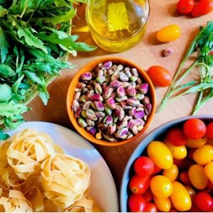Ingredients for arugula pesto pasta. Pistachio nuts in a bowl in the centre, flanked by green arugula leaves, a bottle of olive oil, red and yellow cherry tomatoes and little balls of dry fettuccine