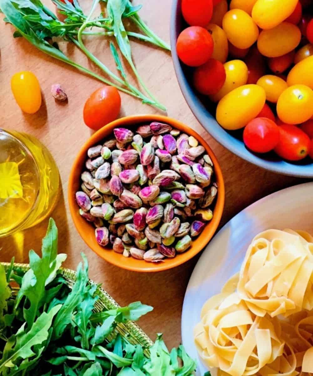 Ingredients for Pasta: Orange bowl of green and pink pistachio nuts, basket of green rocket arugula leaves, plate of dry fettucine, bowl of yellow and red cherry tomatoes, bottle of olive oil