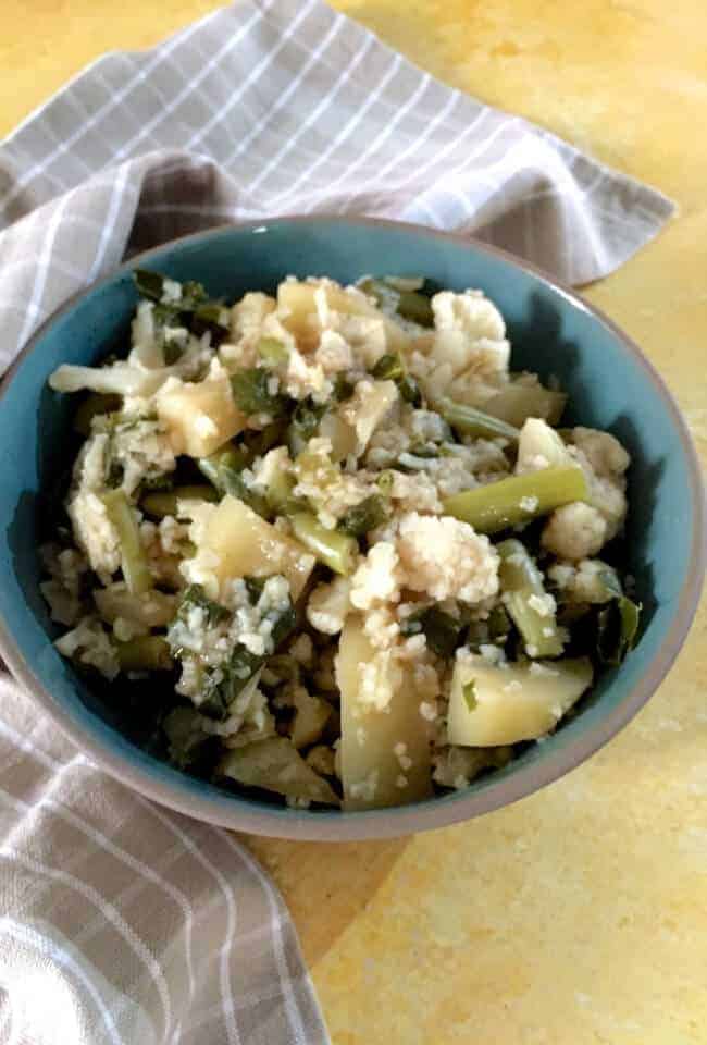 A large bluish green bowl filled with cauliflower stalks, leaves, florets and potatoes and beans boiled to make Mizo Bai, a vegetable stew. A pale brown checked napkin on the left