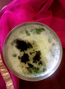Seen from the top, a glass of white buttermilk (diluted and churned yogurt) with agauzy pink fabric as background. Lightly spiced with ginger and with seasoning of black mustard seeds and curry leaves seen floting on top