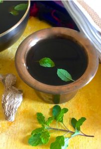 A brass tumbler (traditional cup) with dark jaggery water or panakam, with 2 fresh green tulsi leaves floating. A piece of dry ginger and some more tulsi leaves, the edge of a black bowl and some white fabric are seen around the tumbler