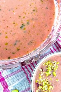 A portion of a glass bowl seen with pink watermelon gazpacho with flecks of green basil oil as garnish. A smaller white bowl with the gazpacho seen to one side and a pink and white checked napkin below