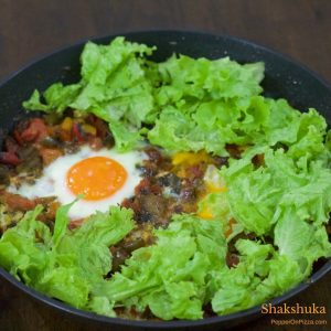 A pan with classic Shakshuka of eggs poached in sauteed tomatoes and red peppers, with a layer of green lettuce arranged along the sides of the pan. Gluten free, easy, delcious