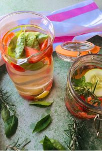 Two glass jars with slices of pink strawberries and with a round slice of cucumber floating on top of one jar. Basil leaves in one and rosemary and mint in the other