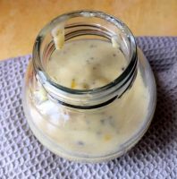 A fat round glass jar half filled with a pale creamy tahini citrus salad dressing, with flecks of black pepper, on a grey beige self checked napkin on a yellow wooden background