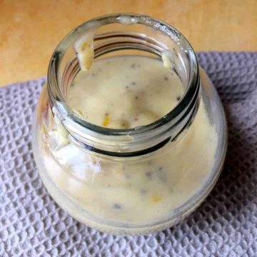 A fat round glass jar half filled with a pale creamy tahini citrus salad dressing, with flecks of black pepper, on a grey beige self checked napkin on a yellow wooden background