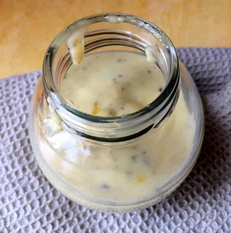 A fat round glass jar half filled with a pale creamy tahini citrus salad dressing, with flecks of black pepper, on a grey beige self checked napkin on a yellow wooden background