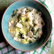 A green bowl with white slices of cooked colocasia, green bits of cabbage and mustard greens in a cloudy liquid. A green and white stirped napkin below and the whloe on a green background