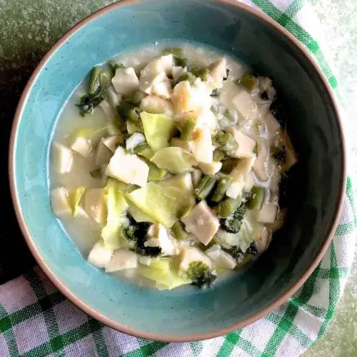 A green bowl with white slices of cooked colocasia, green bits of cabbage and mustard greens in a cloudy liquid. A green and white stirped napkin below and the whloe on a green background