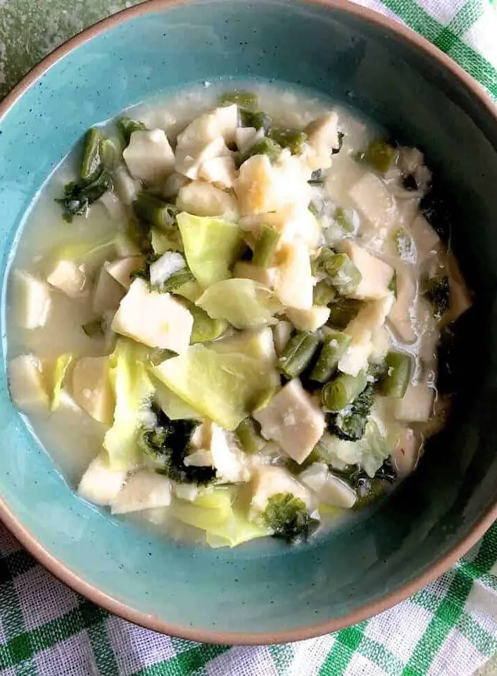 A green bowl with white slices of cooked colocasia, green bits of cabbage and mustard greens in a cloudy liquid. A green and white stirped napkin below and the whloe on a green background