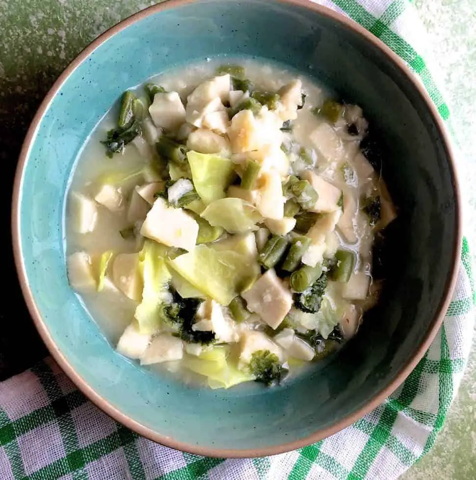 A green bowl with white slices of cooked colocasia, green bits of cabbage and mustard greens in a cloudy liquid. A green and white stirped napkin below and the whloe on a green background