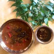 Brass bowl with reddish brown liquid rasam with tomatoes and fried neem flowers seen on the surface. Fresh neem leaves in the background and a brass cup on the side with fried neem flowers. All on a yellow background