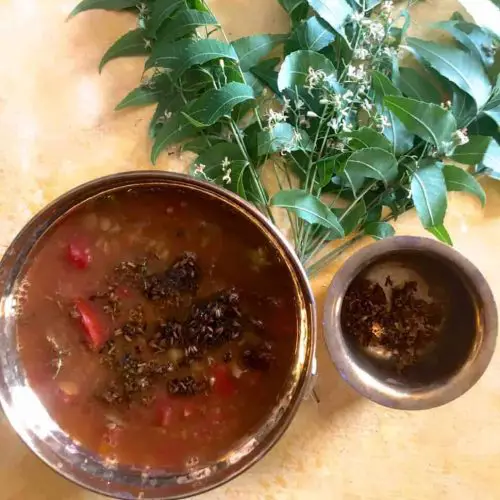 Brass bowl with reddish brown liquid rasam with tomatoes and fried neem flowers seen on the surface. Fresh neem leaves in the background and a brass cup on the side with fried neem flowers. All on a yellow background