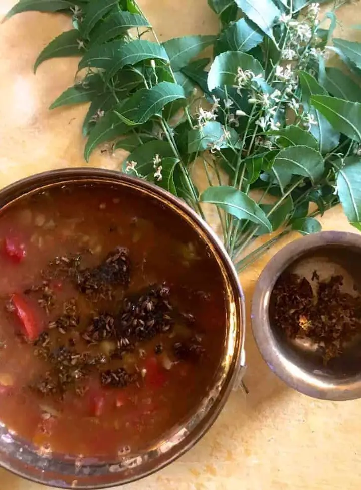 Brass bowl with reddish brown liquid rasam with tomatoes and fried neem flowers seen on the surface. Fresh neem leaves in the background and a brass cup on the side with fried neem flowers. All on a yellow background